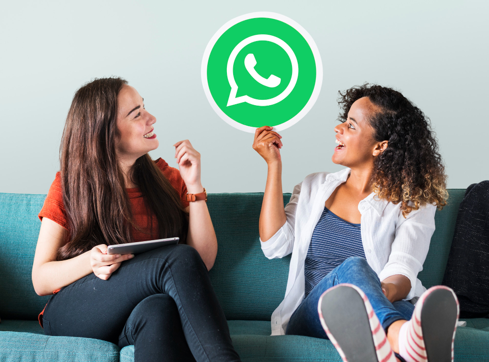 2 mulheres sentadas em um sofá conversando sobre como vender bem pelo WhatsApp Business. A moça da direita segura o simbolo do WhatsApp na mão.